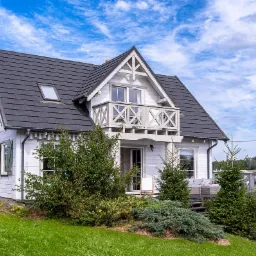 Biały dom z szarą dachówką i balkonem z drewnianą balustradą, otoczony zielenią. Widok z przodu, słoneczny dzień, fragment schodów wejściowych z boku.