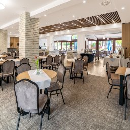 Bobieni Fotografia Wiesław Bobieni - Jasne, przestronne wnętrze restauracji z nakrytymi stołami, krzesłami i dekoracjami kwiatowymi. Widoczne kolumny z cegły i drewniane elementy dekoracyjne.
