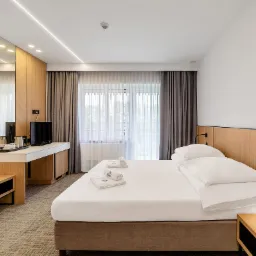 Jasne wnętrze pokoju hotelowego z łóżkiem, biurkiem i telewizorem. Minimalistyczny design, drewniane akcenty, stonowana kolorystyka i delikatne oświetlenie tworzą relaksującą atmosferę.