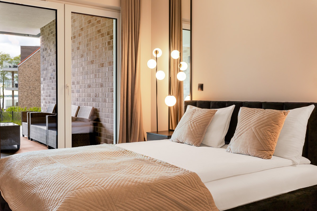 Elegancka sypialnia z wyjściem na balkon: łóżko z beżową narzutą, lampa z kulami, widok na ceglany balkon z meblami ogrodowymi. Stonowana kolorystyka i nowoczesny design.