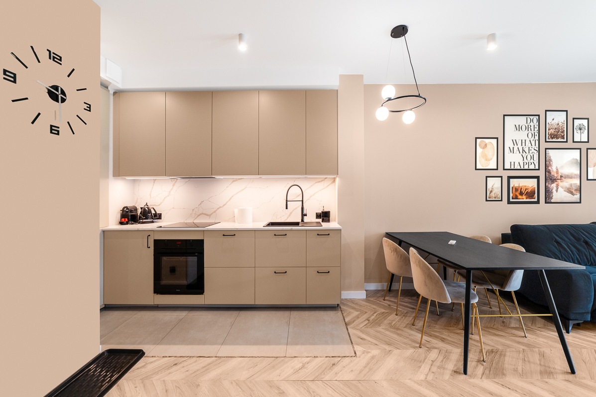 Beżowa kuchnia z marmurowym blatem, czarnym zlewem i piekarnikiem. Nad stołem wisi nowoczesna lampa. Na ścianie galeria obrazków i zegar.