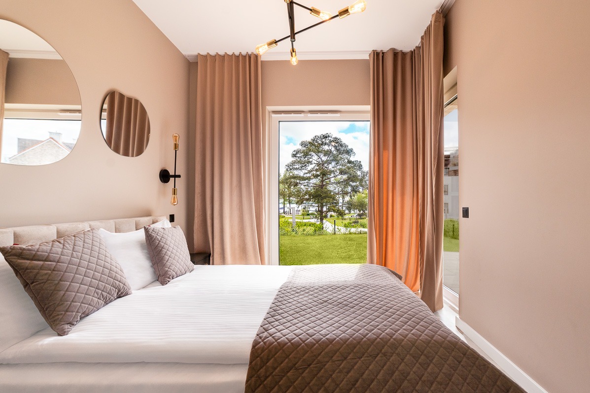 Jasny pokój w Gajewie z łóżkiem, oknem na zieleń i designerskimi lustrami. Lampy w stylu retro. Beżowe ściany i zasłony tworzą przytulną atmosferę.