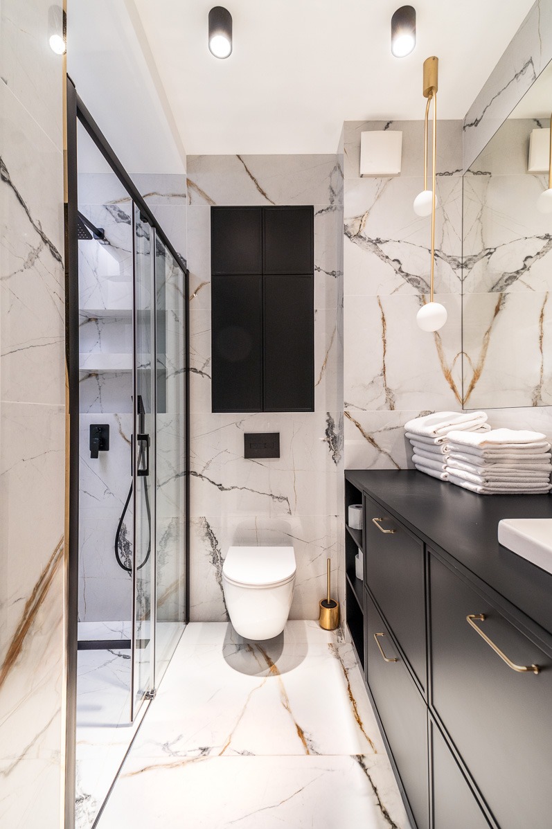 Elegancka łazienka z kabiną prysznicową, toaletą i umywalką na czarnej szafce. Marmurowe płytki, złote akcenty i nowoczesne oświetlenie tworzą spójną całość.