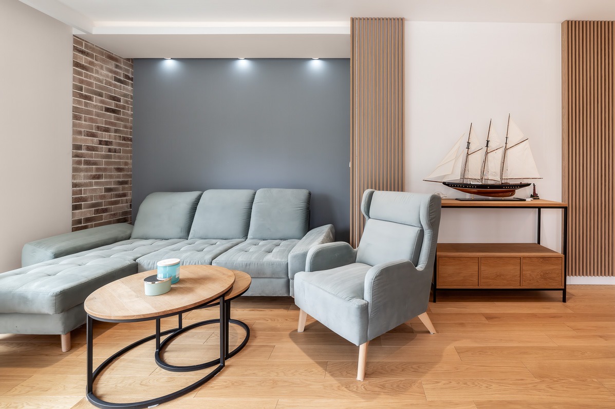 Jasne wnętrze salonu z narożnikiem, fotelem i drewnianym stolikiem kawowym na parkiecie. Na ścianie dekoracyjna cegła i model żaglowca.