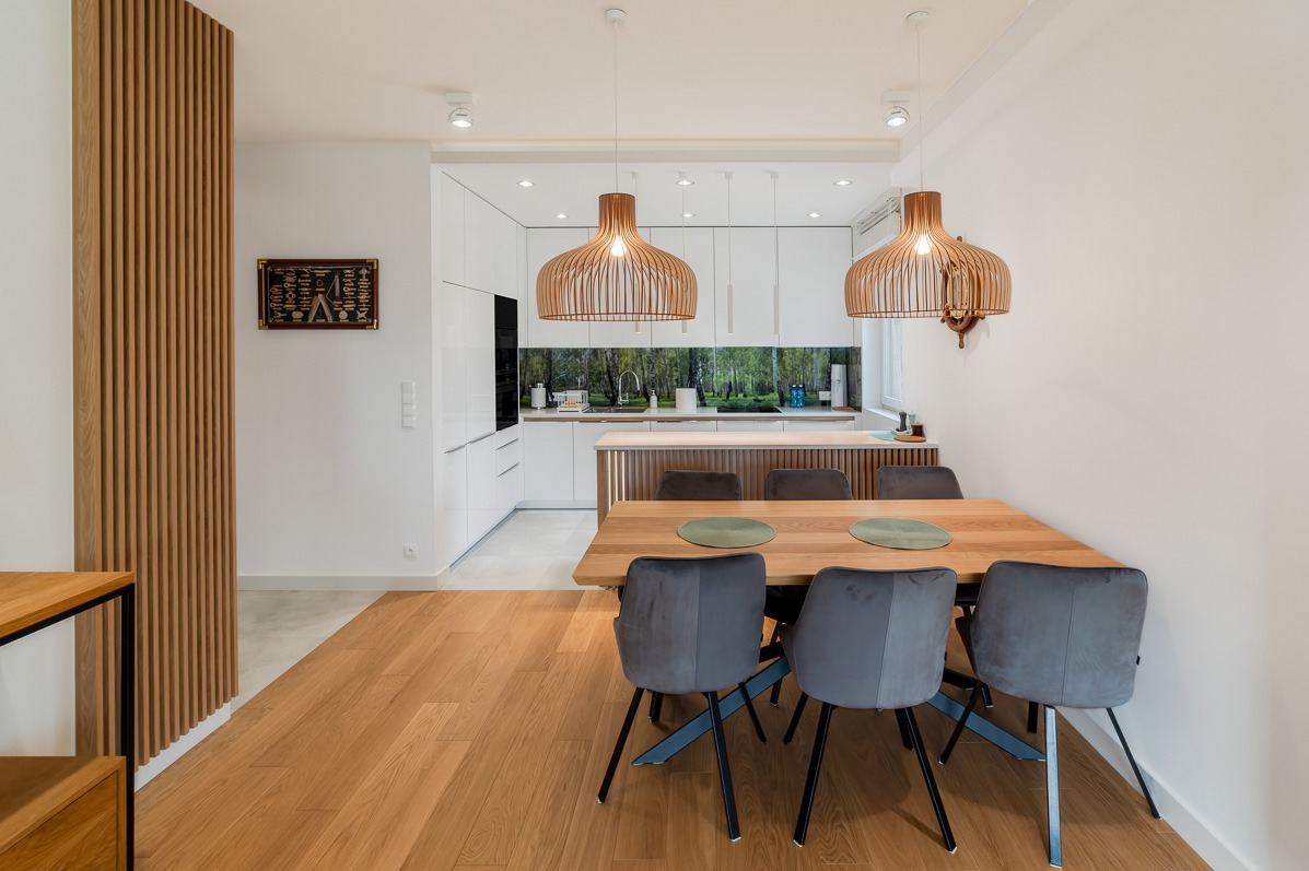 Jasne wnętrze: drewniany stół z szarymi krzesłami i okrągłymi podkładkami, nad nim ażurowe lampy. W tle biała zabudowa kuchenna z leśnym motywem.