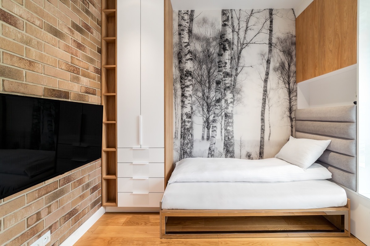 Nowoczesna sypialnia: ceglana ściana z TV, biała szafa, regał z drewna i łóżko z zagłówkiem na tle fototapety z brzozami. Parkiet na podłodze.