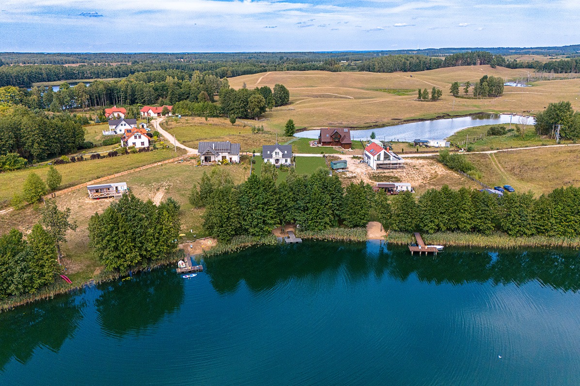 Panoramiczny widok z lotu ptaka na jezioro otoczone domami i lasami, z wyraźnym odbiciem nieba w wodzie i zielonymi brzegami.