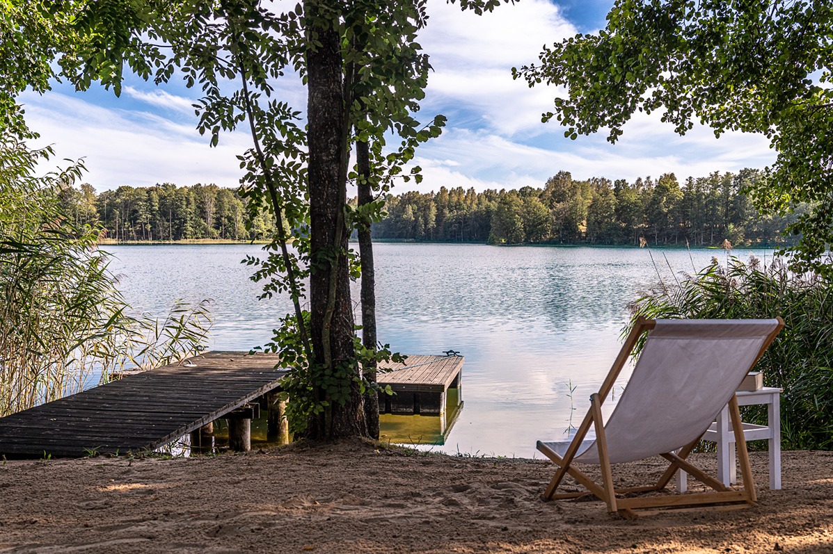 Spokojny krajobraz nad jeziorem z drewnianym pomostem i leżakiem na piaszczystym brzegu, otoczony zielenią drzew i trzcin w słoneczny dzień.