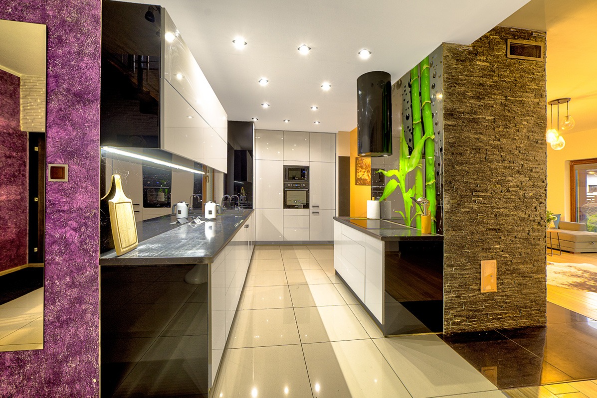 Nowoczesna kuchnia z białymi szafkami, czarnymi blatami i fioletową ścianą. Widoczny fragment salonu, bambusowy motyw na ścianie i oświetlenie punktowe.