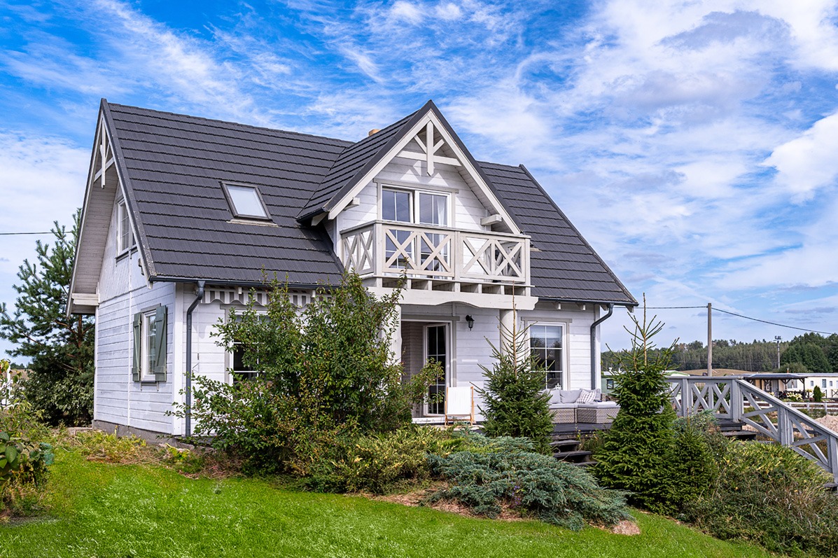 Biały dom z szarą dachówką i balkonem z drewnianą balustradą, otoczony zielenią. Widok z przodu, słoneczny dzień, fragment schodów wejściowych z boku.