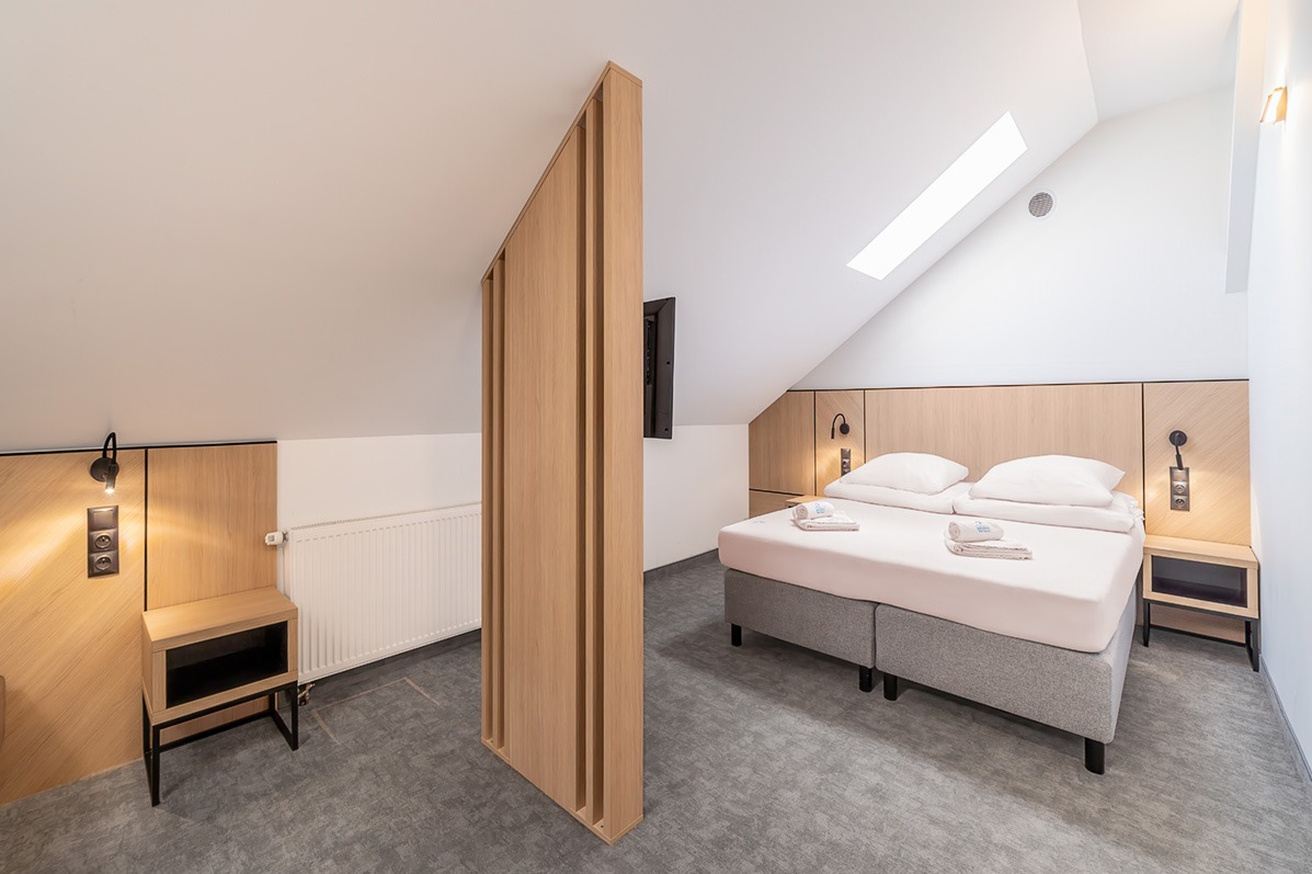 Jasne wnętrze pokoju hotelowego na poddaszu z dwoma łóżkami, drewnianą ścianką działową i minimalistycznym oświetleniem.