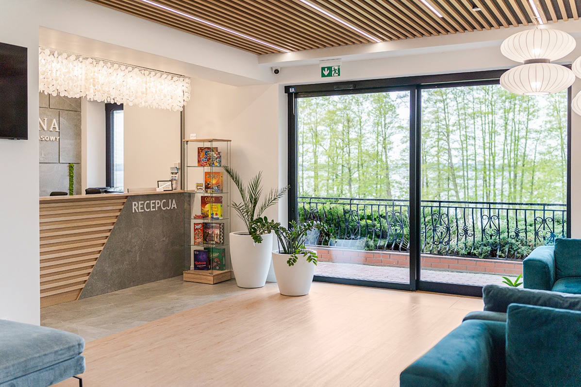 Jasne, przestronne wnętrze recepcji z widokiem na zieleń za oknem. Nowoczesny design łączy drewno i szarość, dopełniają białe donice z roślinami i dekoracyjne oświetlenie.
