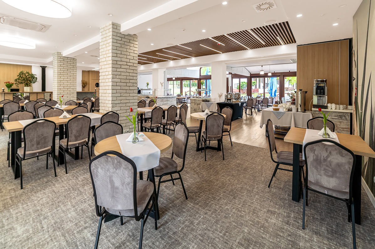 Jasne, przestronne wnętrze restauracji z nakrytymi stołami, krzesłami i dekoracjami kwiatowymi. Widoczne kolumny z cegły i drewniane elementy dekoracyjne.