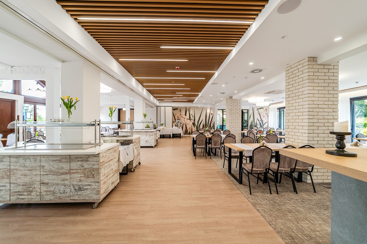 Jasne wnętrze restauracji z drewnianym sufitem, bufetami z kwiatami i nakrytymi stolikami. Ściany zdobi mural z motywem roślinnym.