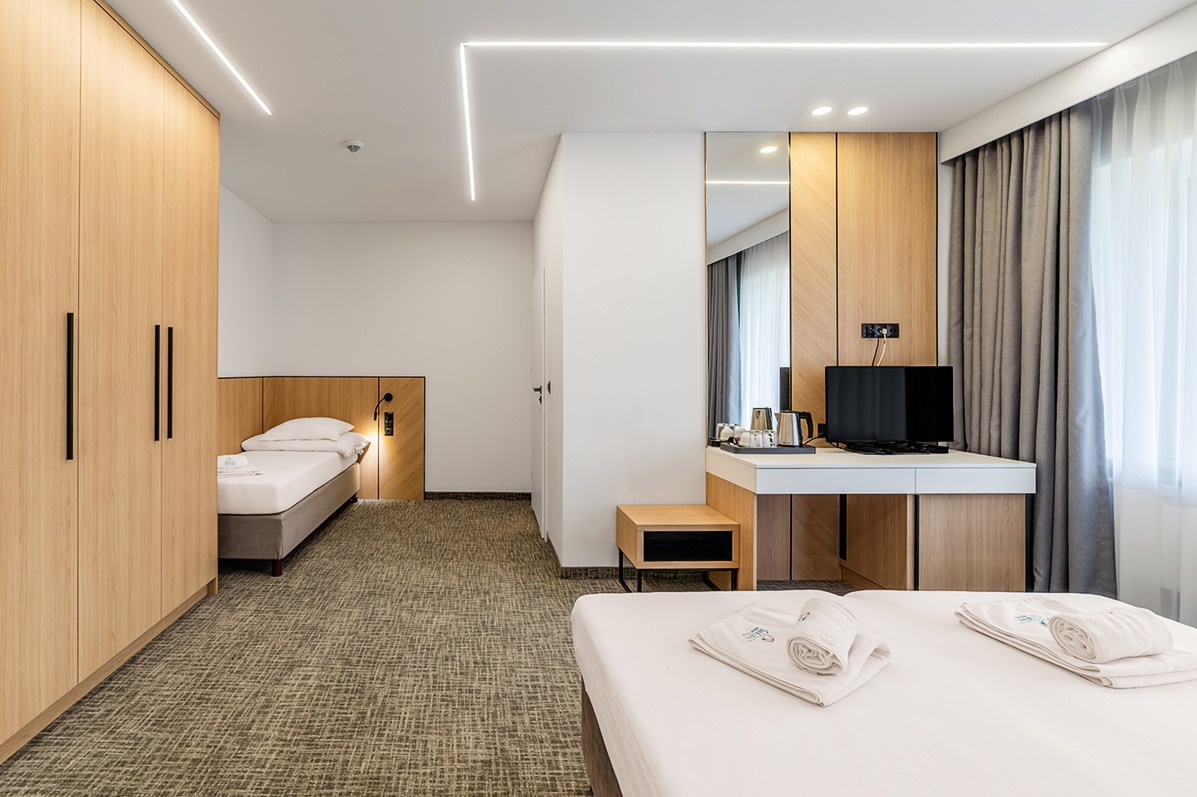 Jasne wnętrze pokoju hotelowego z dwoma łóżkami, biurkiem i szafą. Minimalistyczny design, oświetlenie LED i stonowana kolorystyka tworzą nowoczesny i przytulny klimat.