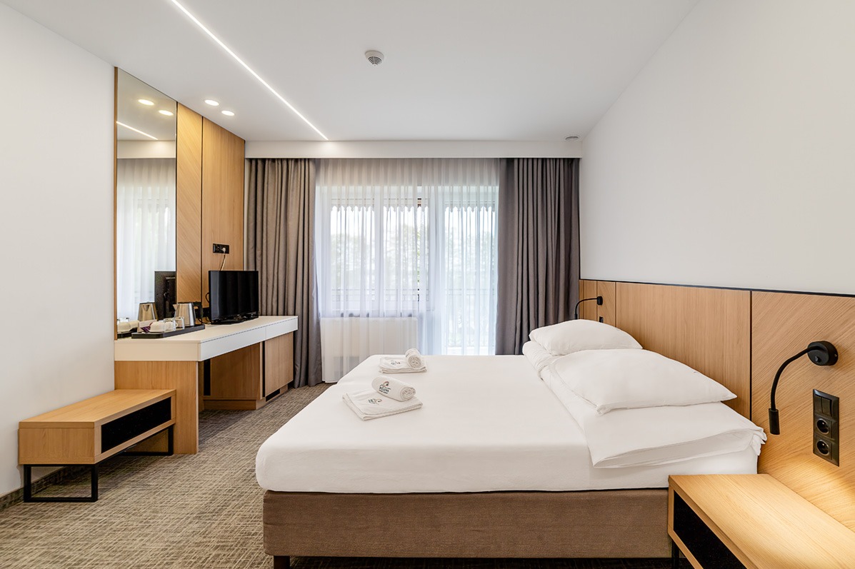Jasne wnętrze pokoju hotelowego z łóżkiem, biurkiem i telewizorem. Minimalistyczny design, drewniane akcenty, stonowana kolorystyka i delikatne oświetlenie tworzą relaksującą atmosferę.