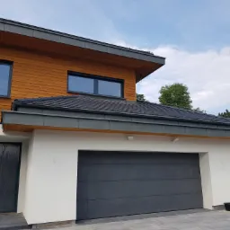 Nowoczesny dom dwukondygnacyjny z drewnianą elewacją na piętrze i białym tynkiem na parterze, z garażem z szarą bramą segmentową i czarnym dachem z orynnowaniem.