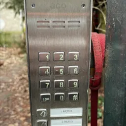 Zewnętrzny panel domofonowy z numerami i przyciskami dzwonków na słupku bramy, widoczne oznaki zużycia na metalowej obudowie i otaczającej konstrukcji.