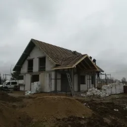 Budowa domu z białych bloczków: wznoszenie więźby dachowej przez ekipę na tle pochmurnego nieba. Widoczne rusztowania, materiały budowlane i teren budowy.