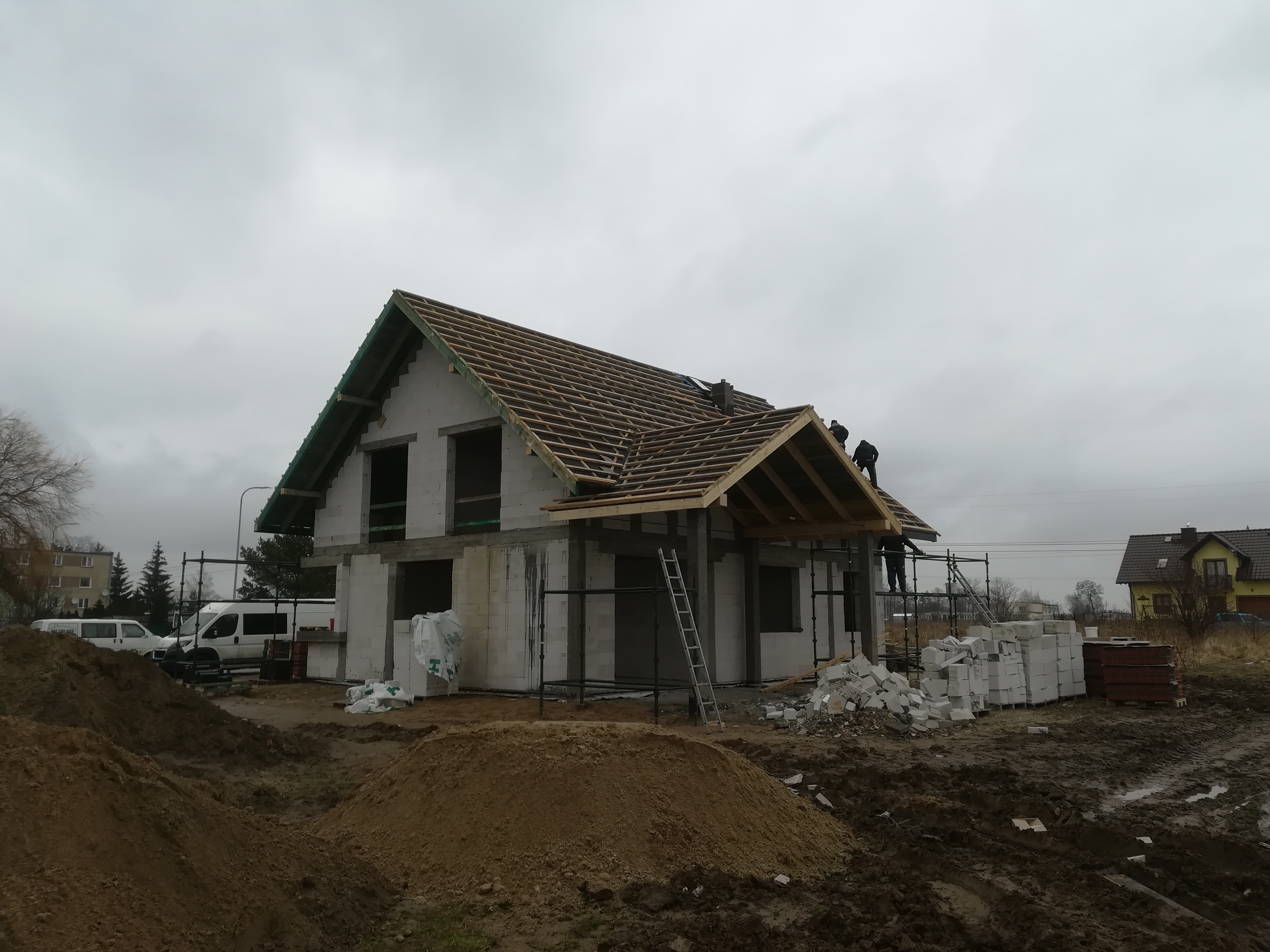 Budowa domu z białych bloczków: wznoszenie więźby dachowej przez ekipę na tle pochmurnego nieba. Widoczne rusztowania, materiały budowlane i teren budowy.