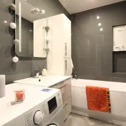 Nowoczesna łazienka w szarościach z białą pralką, umywalką i wanną, ozdobiona pomarańczowym ręcznikiem i świecami na pralce.