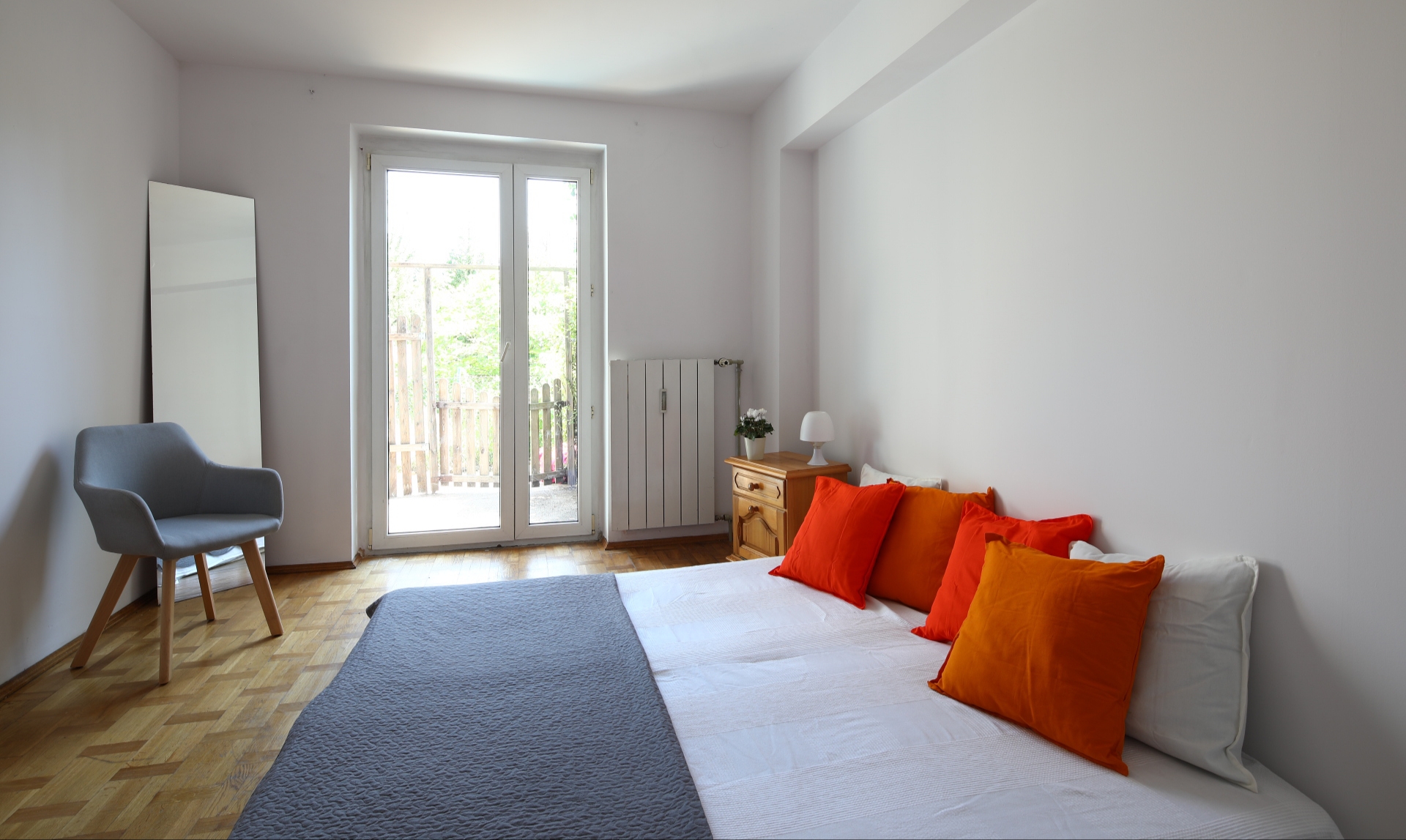 Jasna sypialnia z parkietem, łóżkiem z pomarańczowymi poduszkami, krzesłem i lustrem, z widokiem na balkon przez przeszklone drzwi.