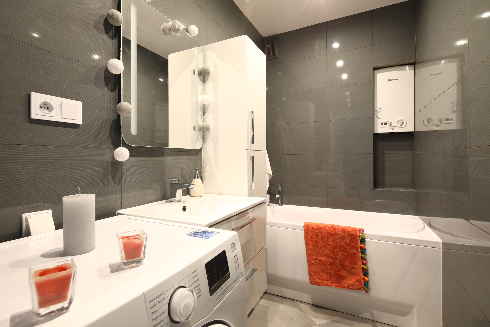 Nowoczesna łazienka w szarościach z białą pralką, umywalką i wanną, ozdobiona pomarańczowym ręcznikiem i świecami na pralce.