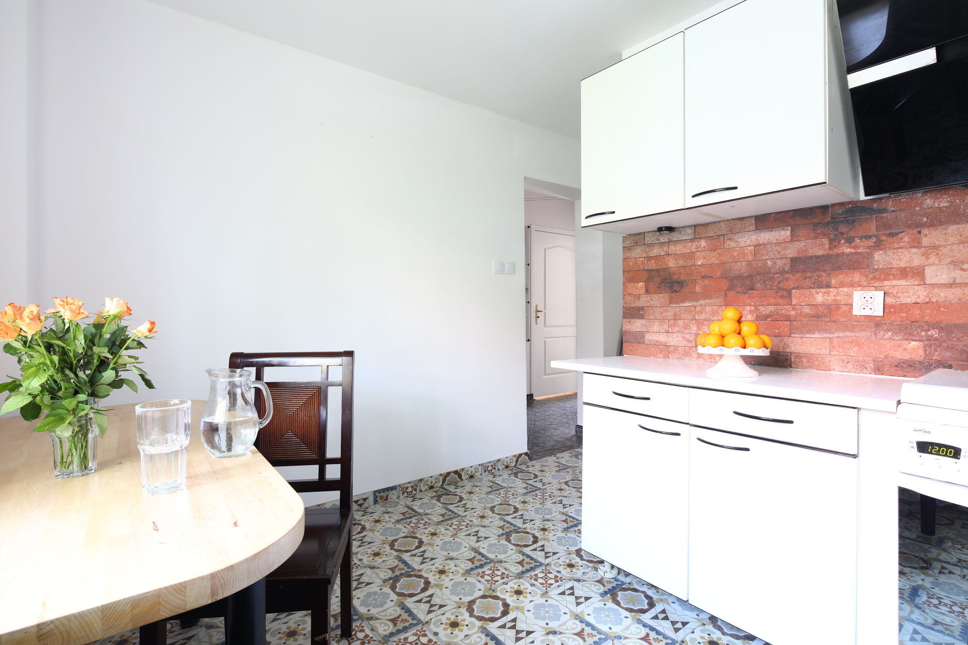 Jasna kuchnia z białymi szafkami, ceglaną ścianą nad blatem, okrągłym drewnianym stołem z bukietem pomarańczowych róż i wzorzystą podłogą.