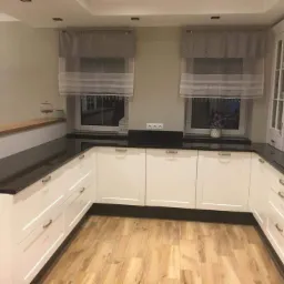 Kuchnia z białymi szafkami i czarnym blatem, widoczne okna z roletami rzymskimi i drewniana podłoga.