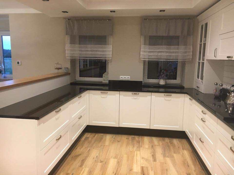 Kuchnia z białymi szafkami i czarnym blatem, widoczne okna z roletami rzymskimi i drewniana podłoga.