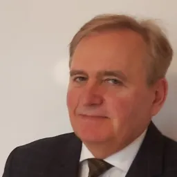 Portret mężczyzny w garniturze i krawacie na jasnym tle, uśmiechający się delikatnie.