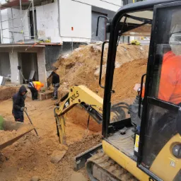 Żółta minikoparka CAT 301.5 w trakcie prac ziemnych na placu budowy domu, widoczni pracownicy w odblaskowych kamizelkach, wykopana ziemia i fragmenty fundamentów.