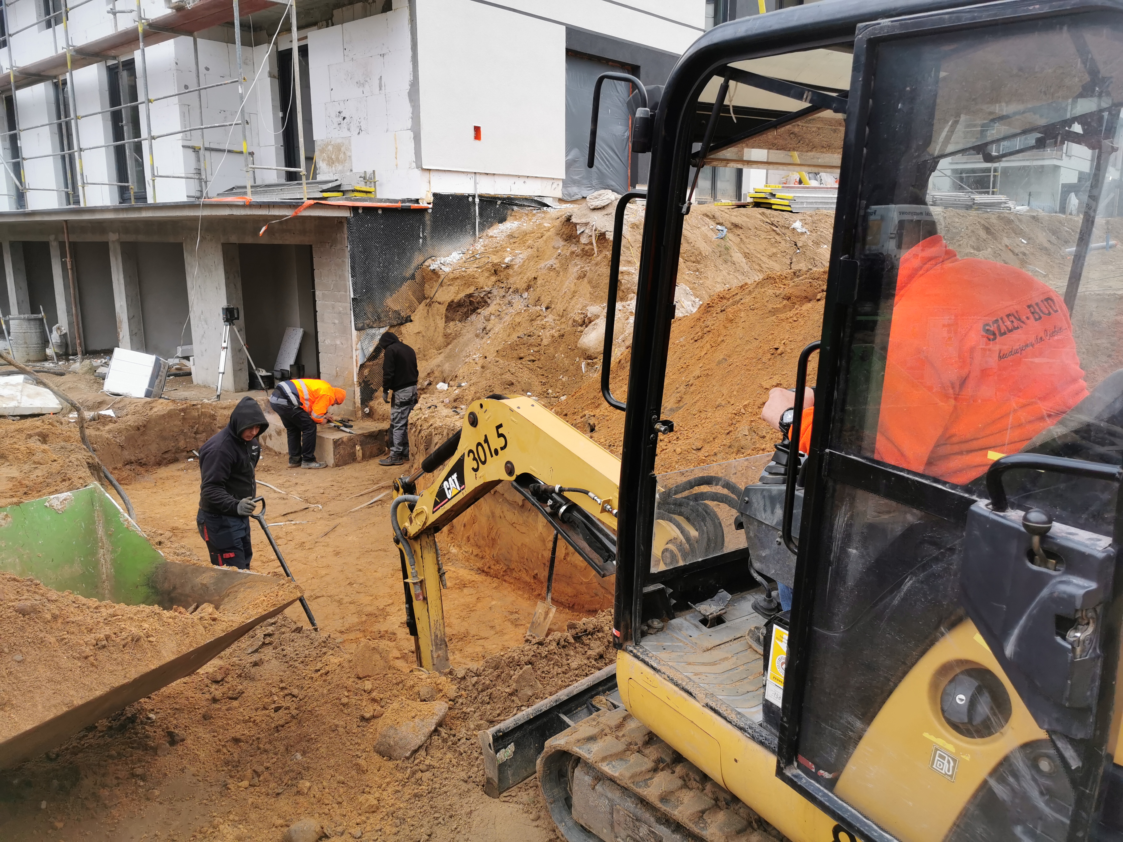 Żółta minikoparka CAT 301.5 w trakcie prac ziemnych na placu budowy domu, widoczni pracownicy w odblaskowych kamizelkach, wykopana ziemia i fragmenty fundamentów.