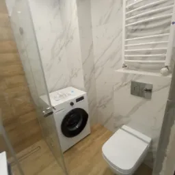Nowoczesna łazienka z marmurowymi płytkami, podwieszaną toaletą, grzejnikiem drabinkowym i pralką, widok na kabinę prysznicową z odpływem liniowym w podłodze.