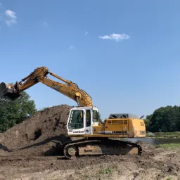 Żółta koparka Liebherr 934 na tle błękitnego nieba i zieleni, nabierająca ziemię z pryzmy, z widocznym zbiornikiem wodnym w tle.