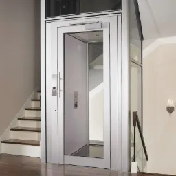 Szklana kabina windy domowej z srebrną ramą, widoczna obok schodów z drewnianymi stopniami i metalową barierką, oświetlona kinkietem na ścianie.