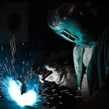 Osoba w masce ochronnej wykonuje prace spawalnicze, widoczne iskry i jasne światło łuku elektrycznego, ciemne tło.