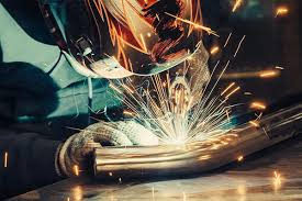 Metalowy element obrabiany iskrzącym się płomieniem, widoczne rękawice ochronne i intensywne światło procesu.