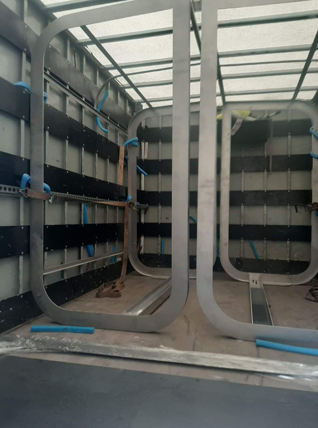 Wnętrze kontenera z metalowymi konstrukcjami w kształcie litery U przymocowanymi do ścian, widoczne niebieskie rurki instalacyjne i pasy mocujące.