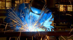 Pracownik w masce ochronnej łączy metalowe elementy za pomocą spawarki, rozbryzg iskier oświetla ciemne otoczenie warsztatu.