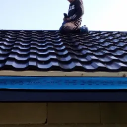 Dekarz na kolanach na nowym, błyszczącym, ciemnym dachu z blachodachówki modułowej. Widoczne detale wykończenia okapu. Niebieska taśma zabezpieczająca.