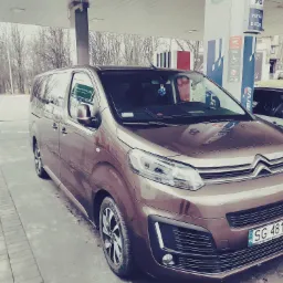 Brązowy van Citroen zaparkowany na stacji benzynowej, widoczny numer rejestracyjny SG 4819T, z przodu laptop na desce rozdzielczej, w tle drzewa bez liści.