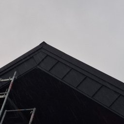 Krajewski Marcin - Ciemny dach z blachy na tle pochmurnego nieba, z widoczną drabiną opartą o krawędź dachu. Ujęcie z dołu, podkreślające geometrię dachu i jego wykończenie.