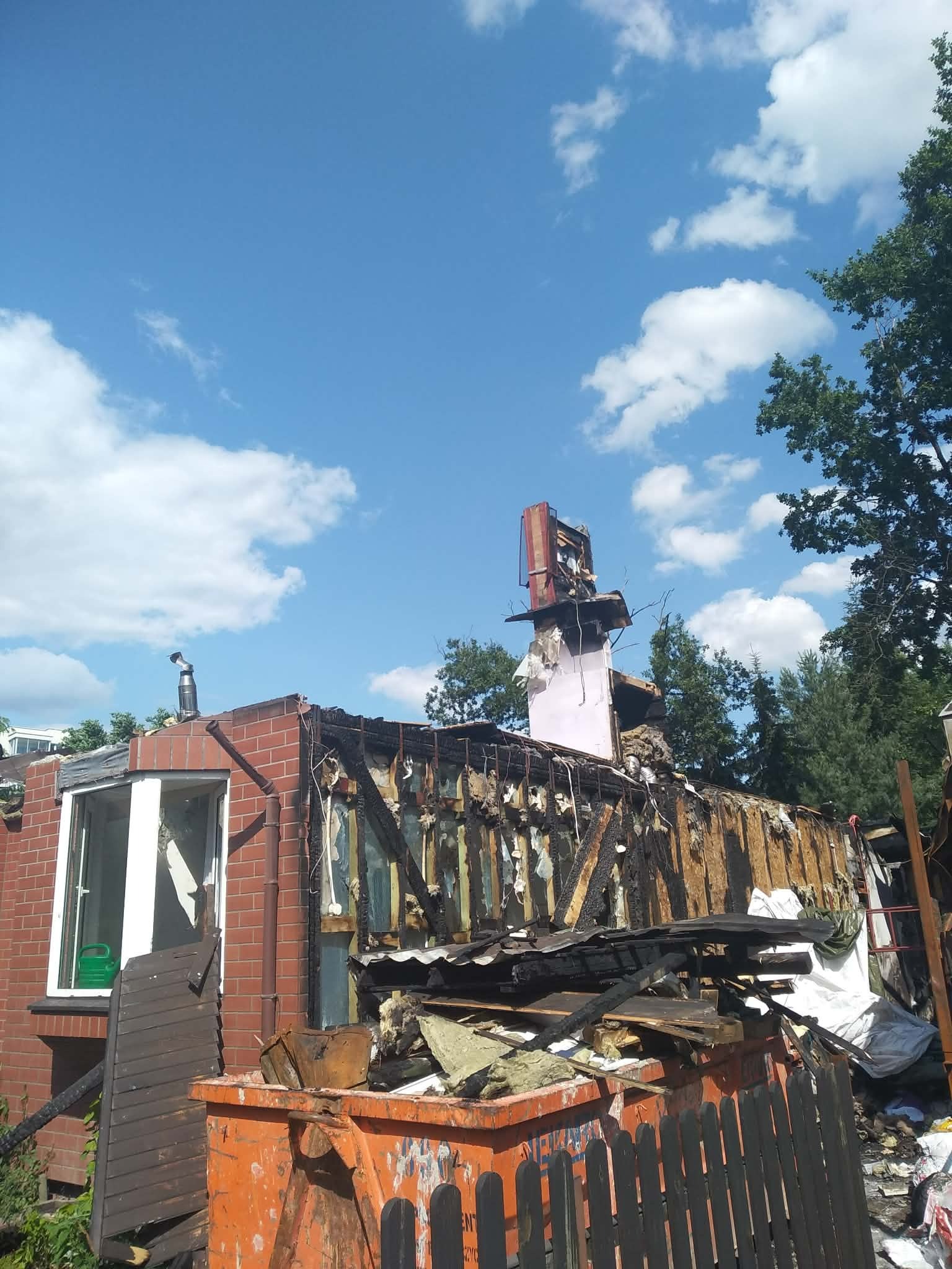 Zniszczony budynek po pożarze z widocznymi spalonymi elementami konstrukcyjnymi i gruzem w kontenerze. Widoczne ceglane ściany i komin. Błękitne niebo z chmurami w tle.