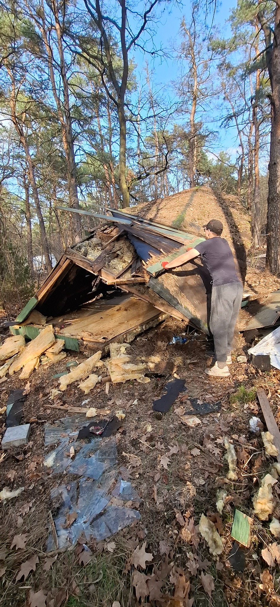 Demontaż zniszczonej konstrukcji dachu przez mężczyznę w lesie. Widoczne fragmenty dachu, izolacja i rozbite szkło na ziemi.