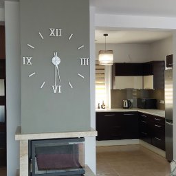 MÓJ SZEŚCIAN - Salon z kominkiem wykończonym marmurem, nad nim duży zegar ścienny z cyframi rzymskimi, w tle widoczna kuchnia z ciemnymi szafkami i jasnym blatem.
