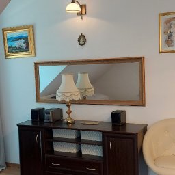 MÓJ SZEŚCIAN - Ciemny drewniany kredens z lustrem w złotej ramie, lampą stołową z frędzlami, głośnikami i ozdobnymi koszykami, uzupełniony obrazami w złotych ramach i beżowym krzesłem na tle białej ściany.