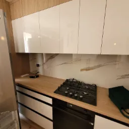 Nowoczesna kuchnia z białymi szafkami wiszącymi, drewnianym blatem, płytą gazową i piekarnikiem, uzupełniona o lodówkę z 10-letnią gwarancją i marmurową ścianę.