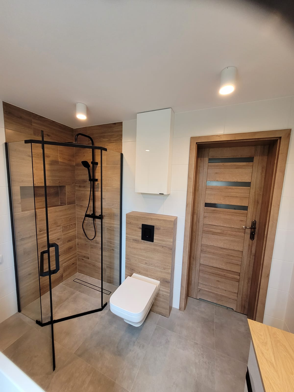 Nowoczesna łazienka z kabiną prysznicową typu walk-in z czarnymi ramami, wiszącą toaletą z drewnopodobną obudową i drewnianymi drzwiami z ciemnymi intarsjami, oświetlona punktowymi lampami.