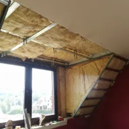 Ocieplenie poddasza wełną mineralną, widoczne metalowe profile konstrukcyjne pod płyty karton-gips, okno z widokiem na domy w tle.